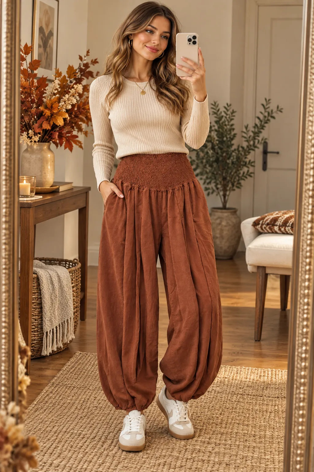 A woman takes a mirror selfie in a cozy room, wearing a cream ribbed long-sleeve top and high-waisted rust-brown corduroy harem pants with white sneakers.