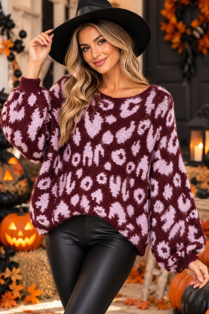 A woman wearing a fuzzy knit sweater with a burgundy and light pink abstract animal print, black faux-leather pants, and a black wide-brim hat, posed in a Halloween-themed setting with pumpkins and autumn decorations.