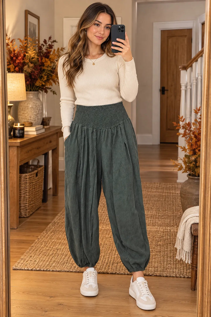 A woman takes a mirror selfie in a home hallway wearing a cream long-sleeve top, dark green high-waisted balloon pants, and white sneakers.