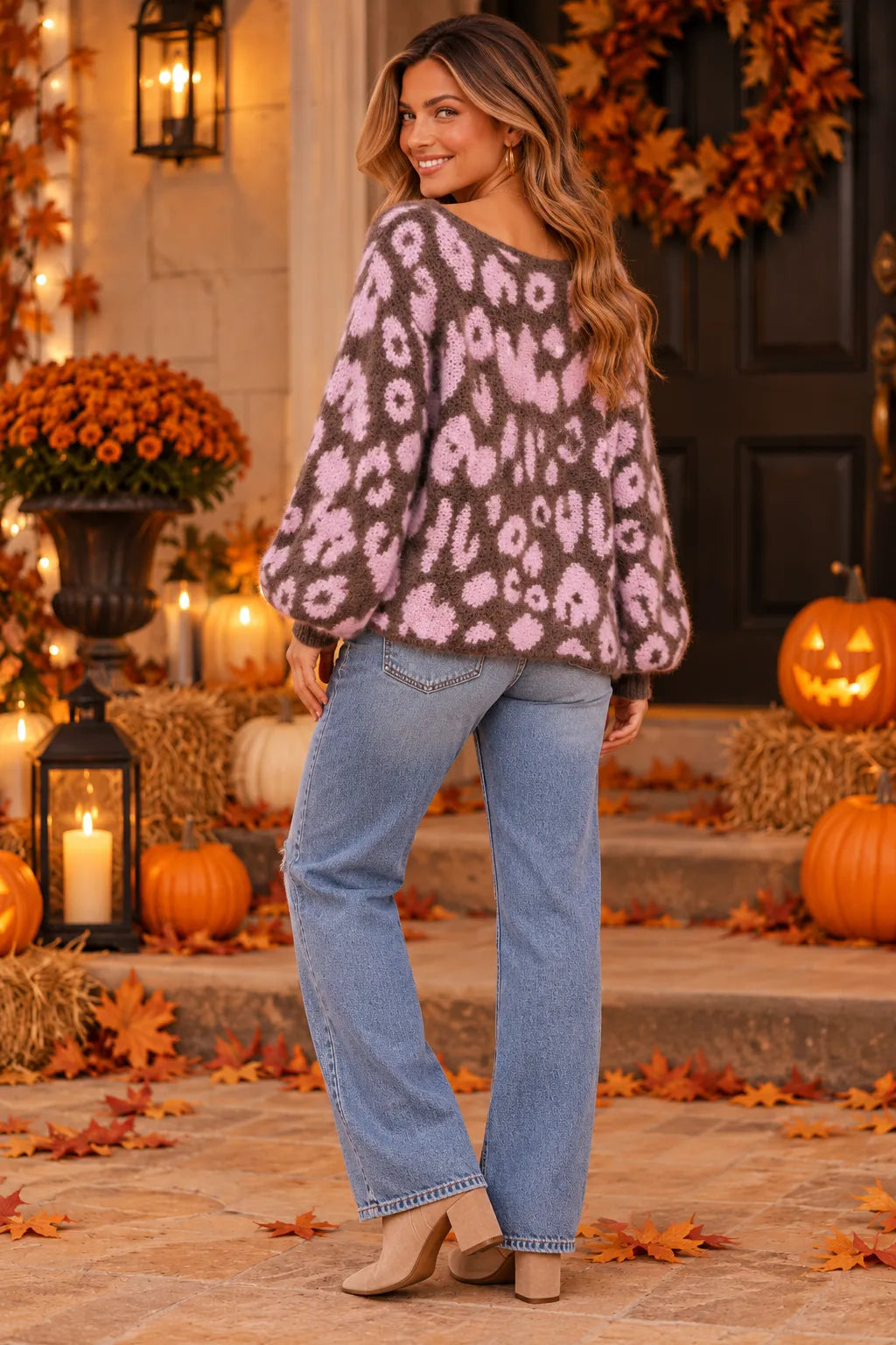 A woman stands on a porch wearing a fuzzy knit sweater with a pink and brown animal print, paired with light blue jeans and beige heels. The background features autumn decorations including pumpkins, fall leaves, a wreath, and warm lanterns.