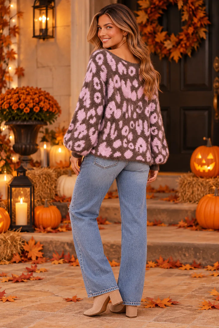 A woman stands on a porch wearing a fuzzy knit sweater with a pink and brown animal print, paired with light blue jeans and beige heels. The background features autumn decorations including pumpkins, fall leaves, a wreath, and warm lanterns.