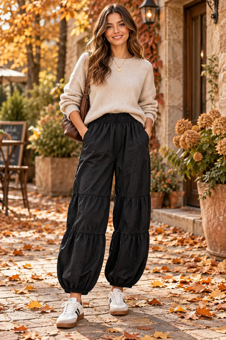 A woman stands outdoors on a stone path covered with fallen leaves, wearing a cream knit sweater, black tiered wide-leg pants, and white sneakers. She is posed in front of a rustic building with plants and autumn foliage.