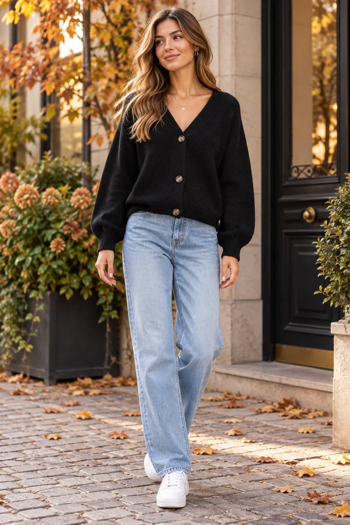 A woman walking outdoors on a cobblestone path wearing a black button-front cardigan, light-wash blue jeans, and white sneakers, with autumn plants and a building entrance in the background.