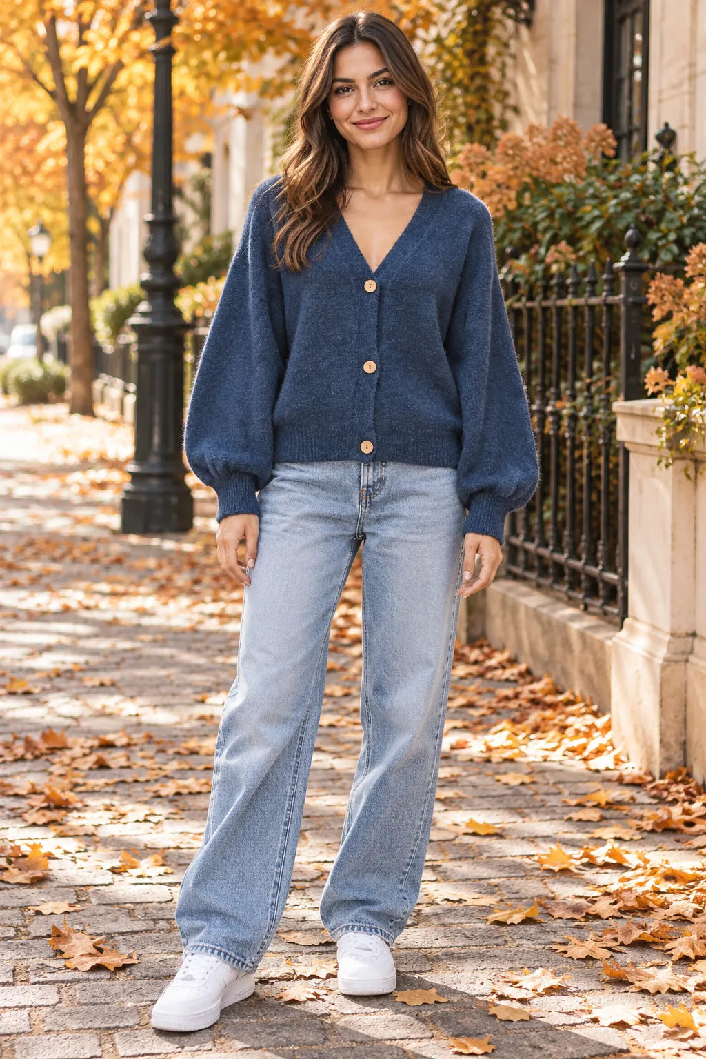 A woman stands on a leaf-strewn brick sidewalk wearing a navy blue knit cardigan with light-wash wide-leg jeans and white sneakers, set against an autumnal street background.