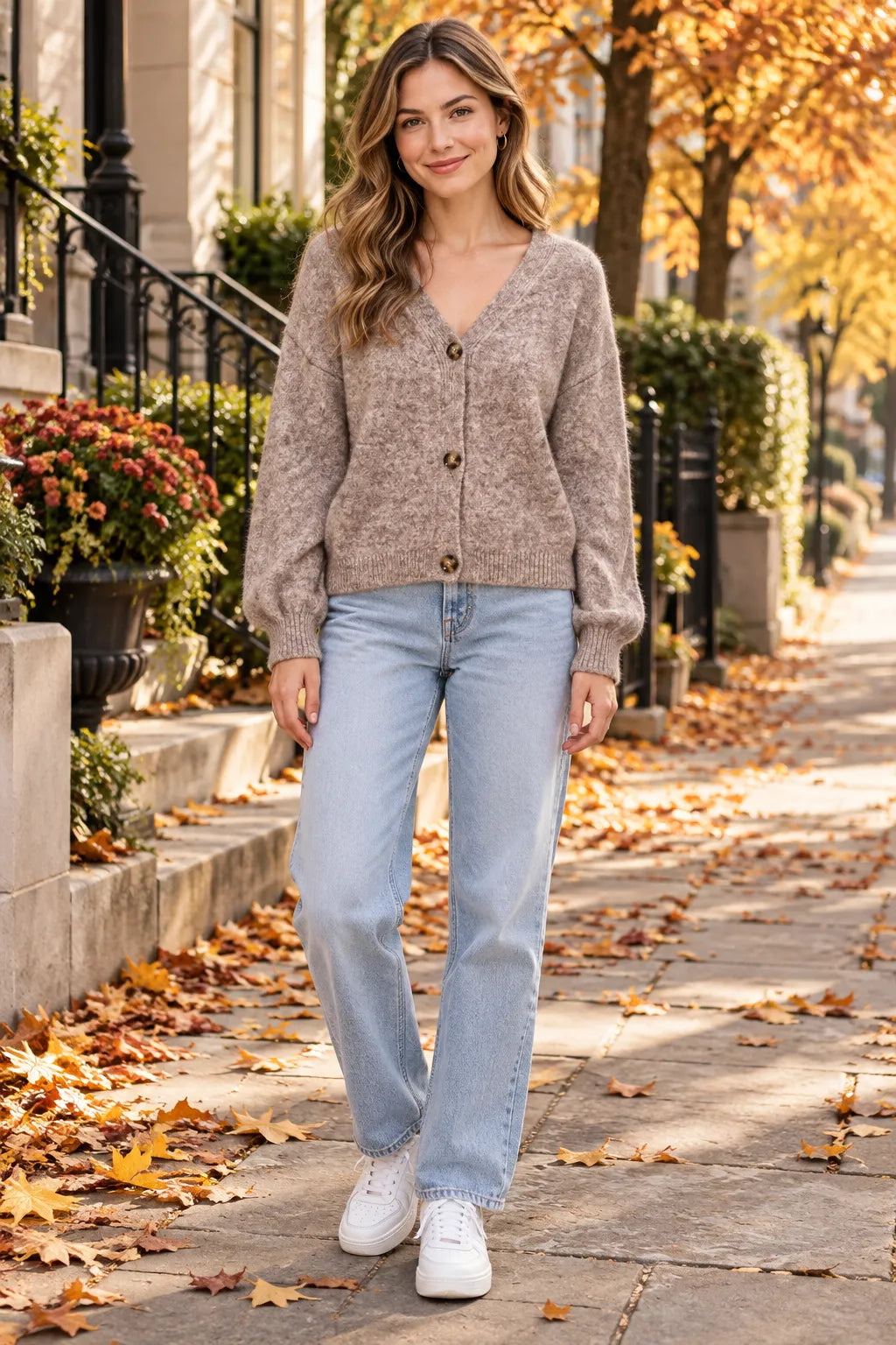 A woman stands on an autumn city sidewalk wearing a taupe knit button-front cardigan, light-wash blue jeans, and white sneakers.
