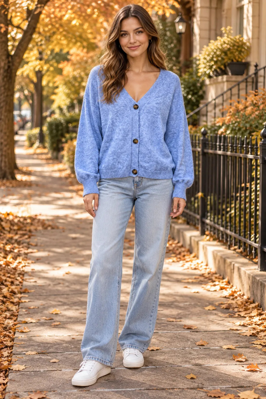 A woman stands on an autumn leaf-covered sidewalk wearing a light blue marled cardigan, light-wash wide-leg jeans, and white sneakers.