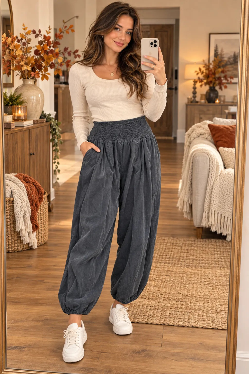 A woman takes a mirror selfie in a cozy living room, wearing dark gray high-waisted balloon pants with a smocked waistband, a cream long-sleeve top, and white sneakers.