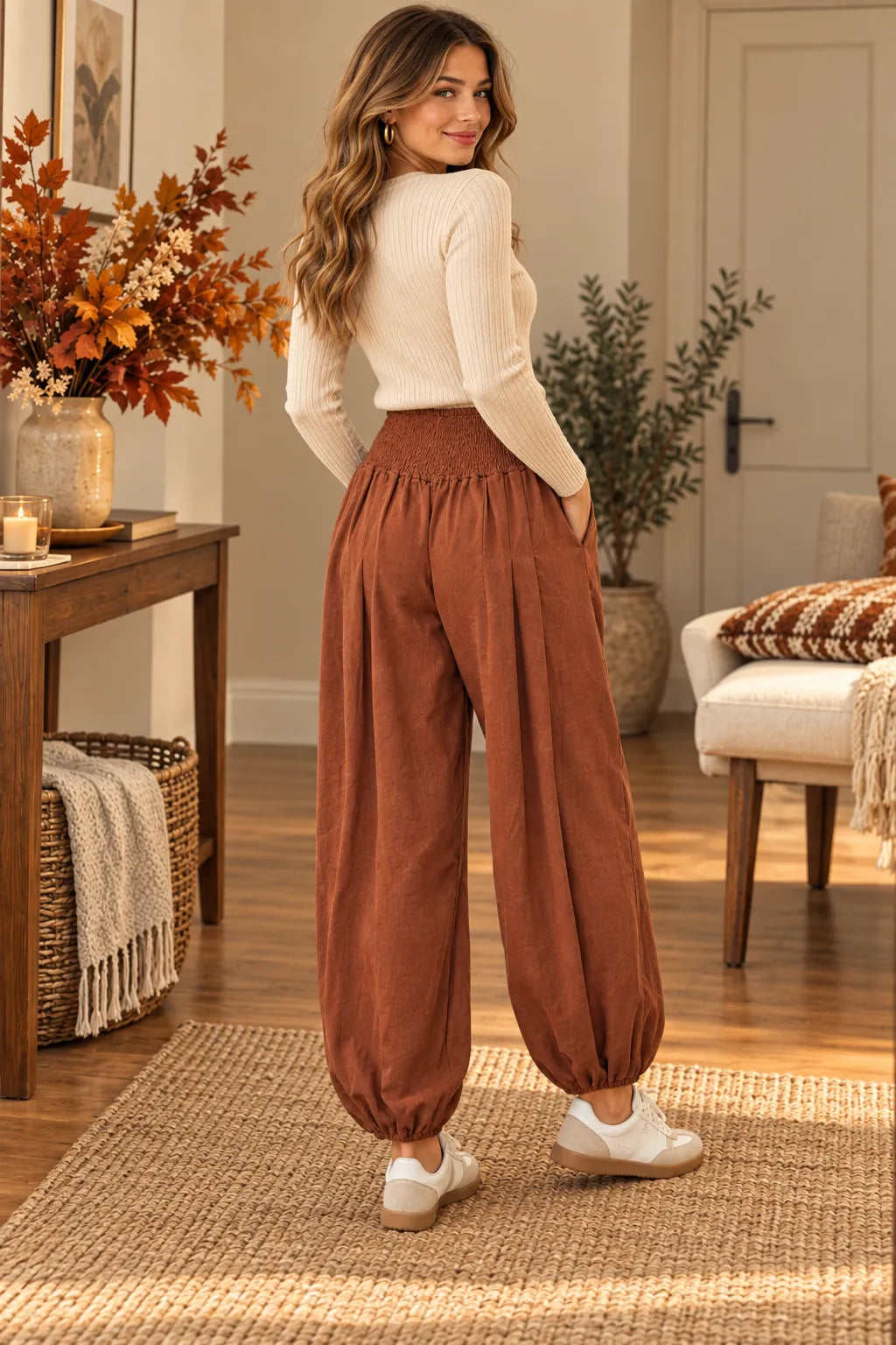 A woman stands in a warmly lit living room wearing loose, high-waisted rust-brown harem pants with a cream ribbed long-sleeve top and white sneakers.