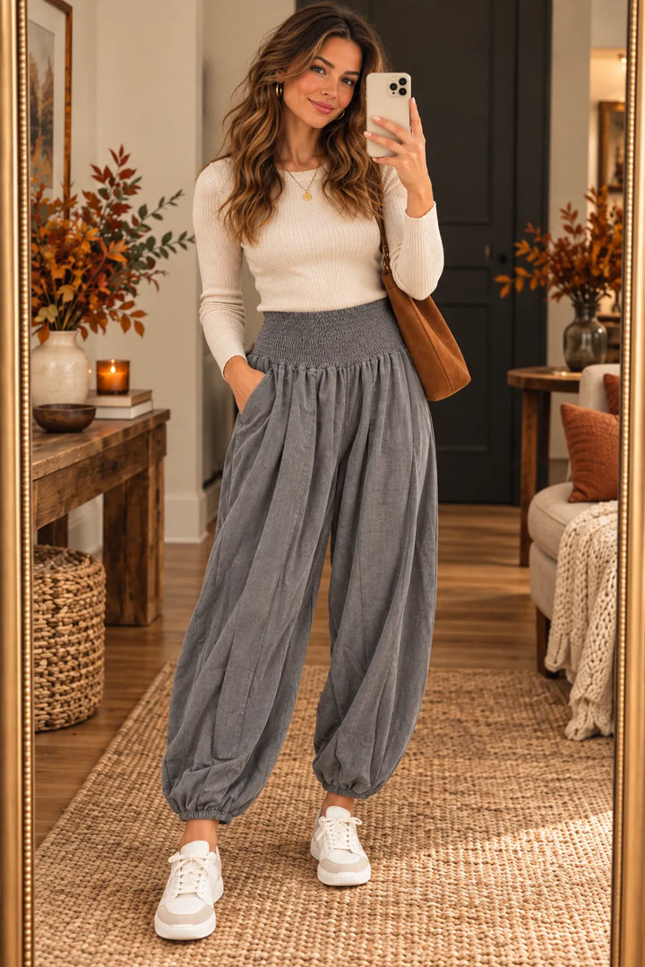 A woman takes a mirror selfie in a cozy room, wearing a cream ribbed long-sleeve top, high-waisted gray harem-style pants, and white sneakers, with a brown shoulder bag.
