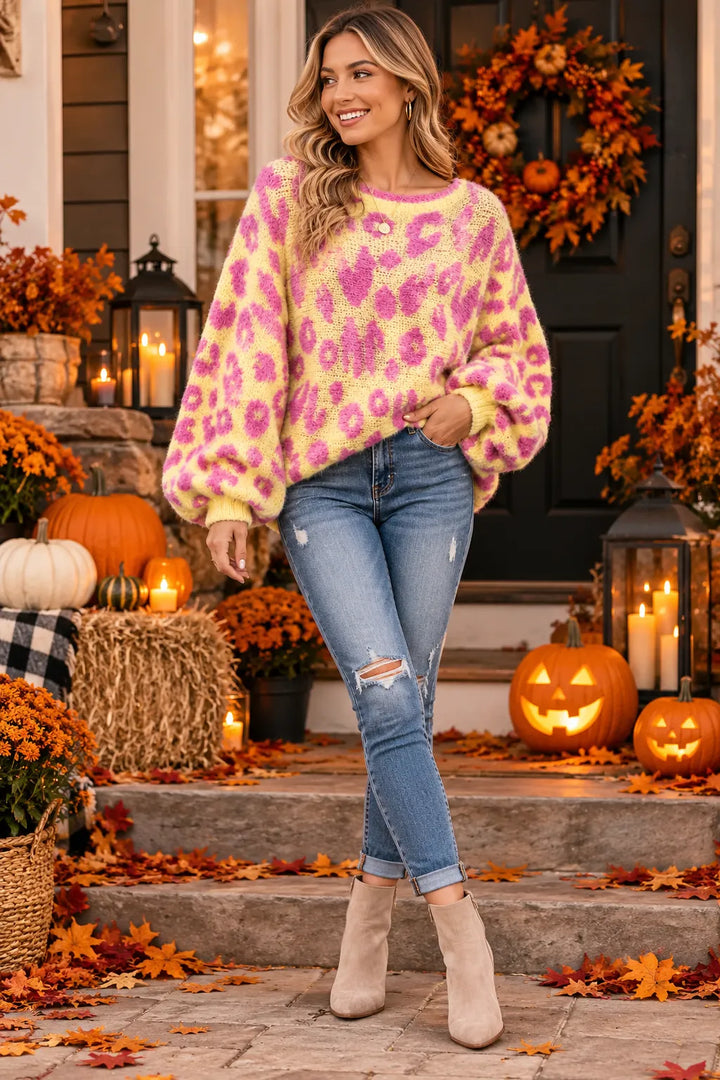 A woman stands on front steps wearing a fuzzy knit sweater with a pink and yellow leopard print, blue jeans, and beige ankle boots. The background shows an autumn-themed porch with pumpkins, fall leaves, lanterns, and a wreath.