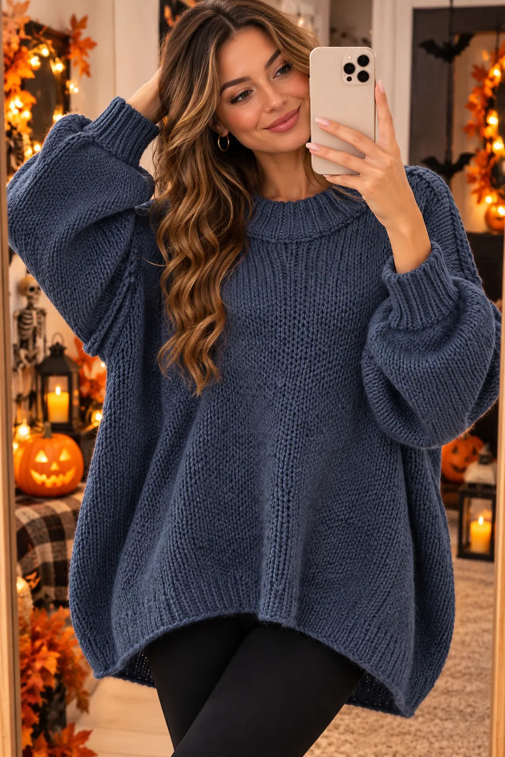 A woman takes a mirror selfie wearing a chunky, oversized knit sweater in a muted slate blue color. The background is a cozy, warmly lit Halloween-themed room with pumpkins, autumn leaves, and string lights.
