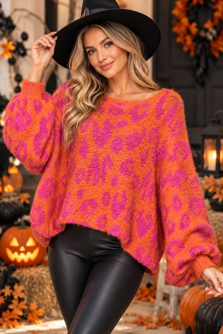 A woman wearing a fuzzy knit sweater with a bright orange and hot pink leopard-like pattern, black pants, and a black witch hat, posed in a Halloween-themed setting with pumpkins and autumn decorations.
