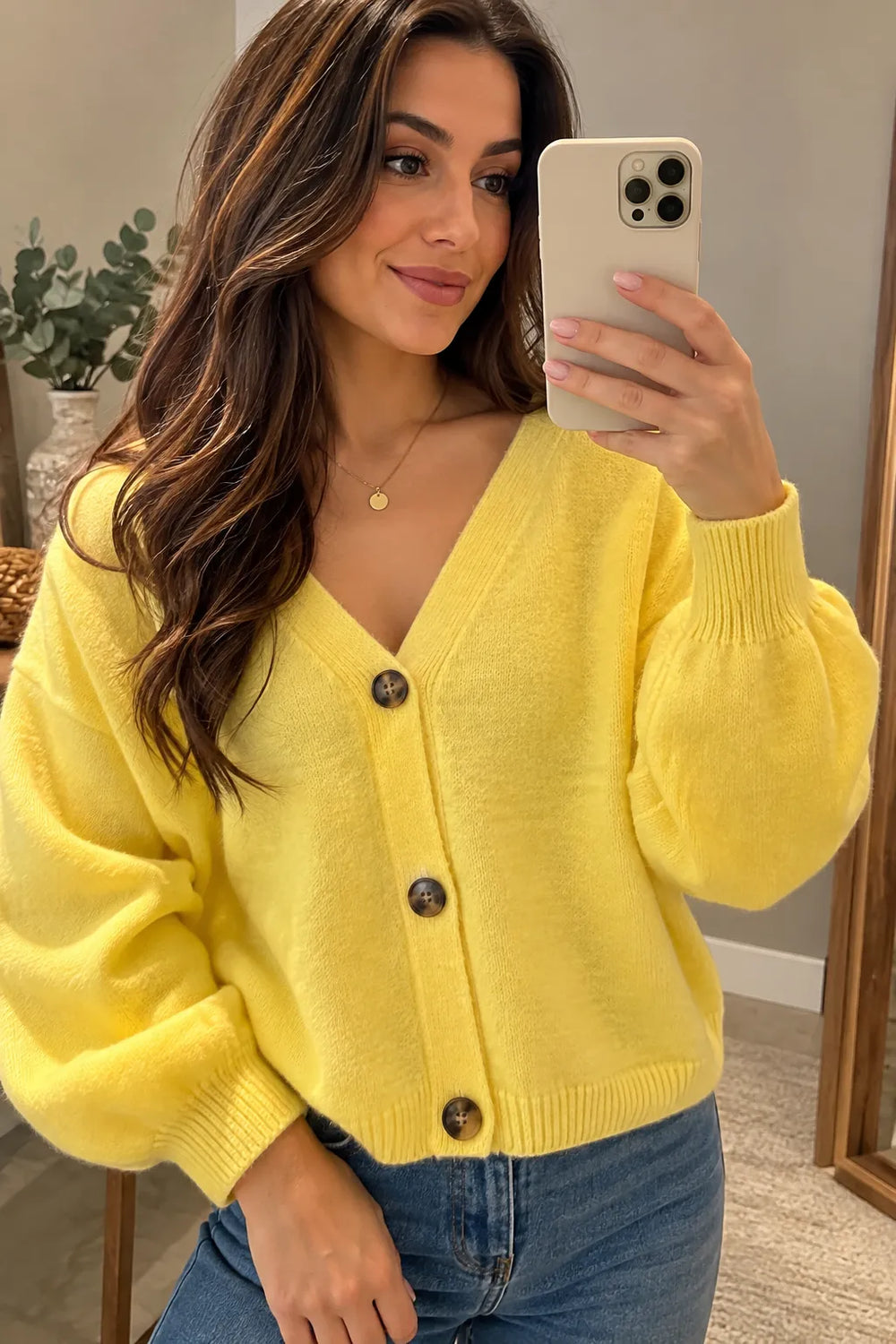 A woman takes a mirror selfie wearing a bright yellow knit cardigan sweater with dark brown buttons and blue jeans.