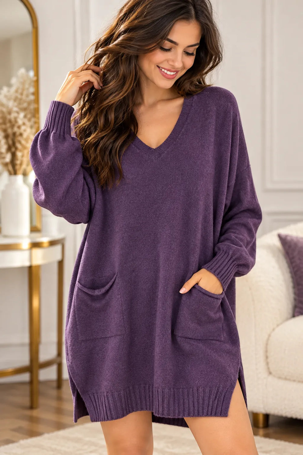A woman wearing an oversized purple knit sweater dress with a V-neck, long sleeves, and front patch pockets, standing in a bright living room.