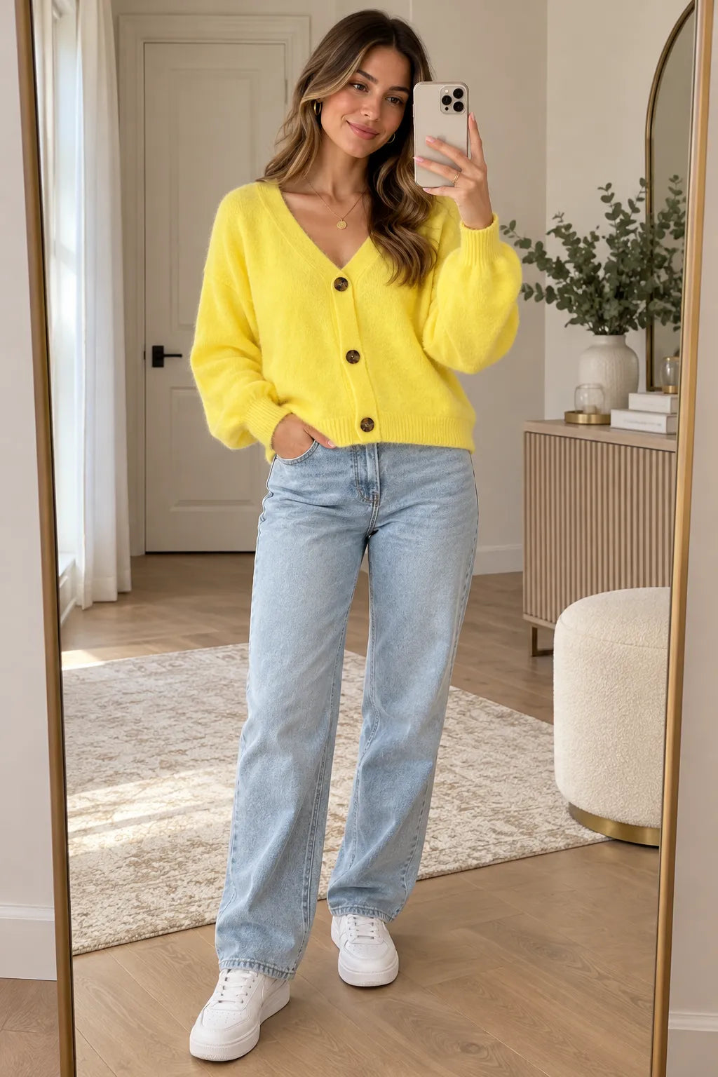A woman takes a mirror selfie in a bright room, wearing a fuzzy bright yellow cardigan with dark buttons, light-wash blue jeans, and white sneakers.