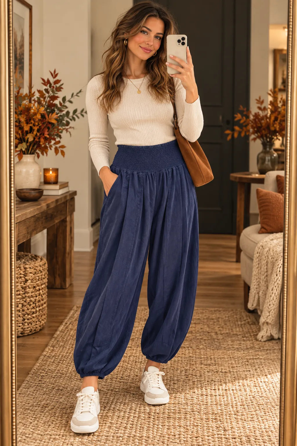 A woman takes a mirror selfie in a cozy room, wearing a cream ribbed long-sleeve top, navy blue high-waisted balloon pants, and white sneakers, with a brown shoulder bag.