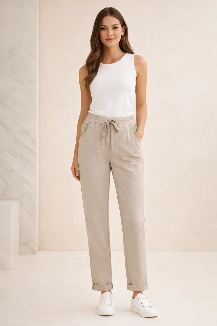 Woman wearing a white sleeveless top and beige pants against a plain background