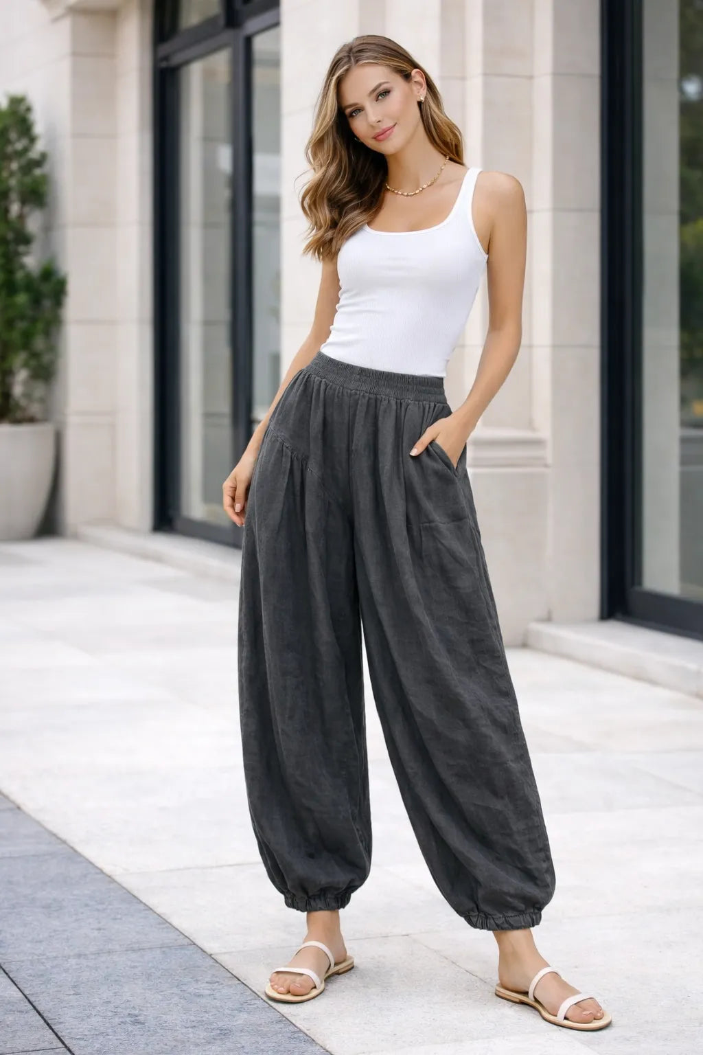 Woman wearing a white tank top and dark gray wide-leg pants standing in front of a building.