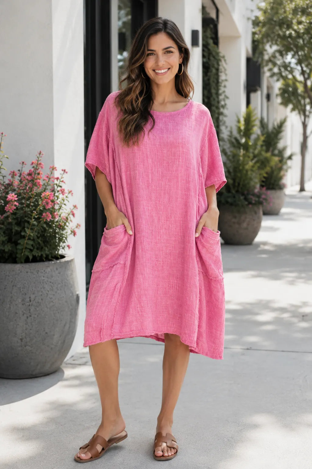 A woman wearing a loose, short-sleeve pink linen-blend dress with large front patch pockets, standing outdoors on a sunlit sidewalk next to a white building.