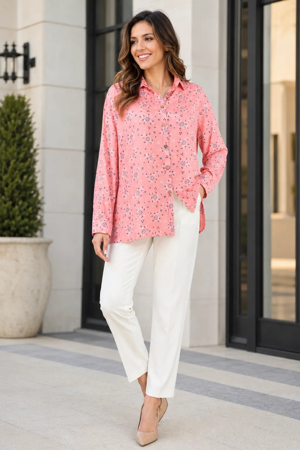 Woman wearing a pink floral blouse and white pants standing outside a building.