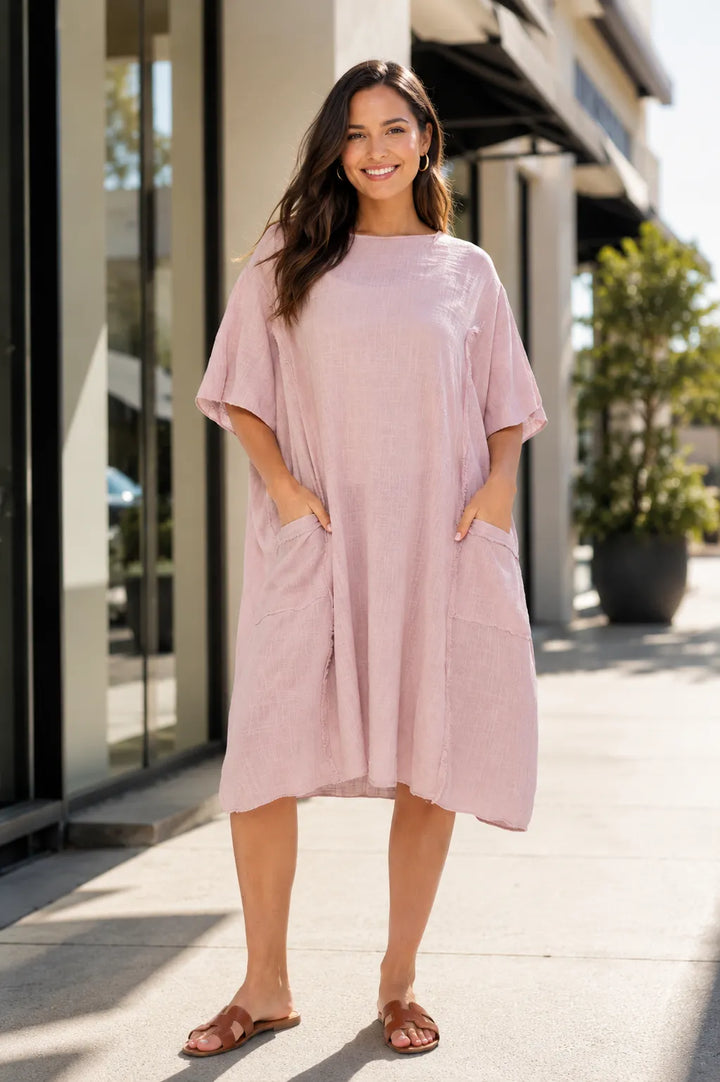 A woman wearing a loose, short-sleeve midi dress in a dusty pink color, standing on a sunny sidewalk in front of a modern building.