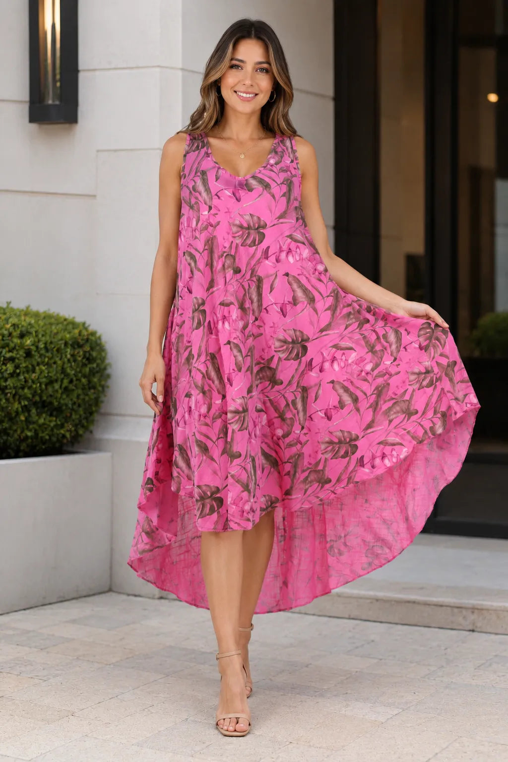 A woman wearing a sleeveless high-low midi dress in bright pink with a brown tropical floral print, standing outdoors in front of a modern building entrance.