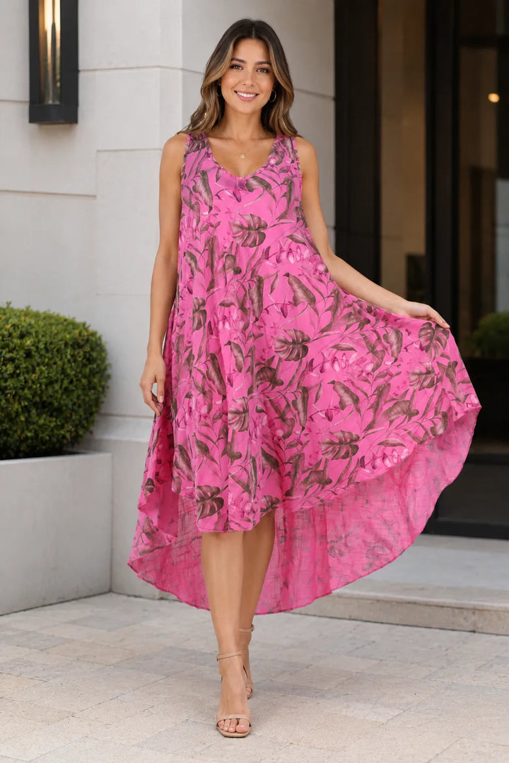 A woman wearing a sleeveless high-low midi dress in bright pink with a brown tropical floral print, standing outdoors in front of a modern building entrance.