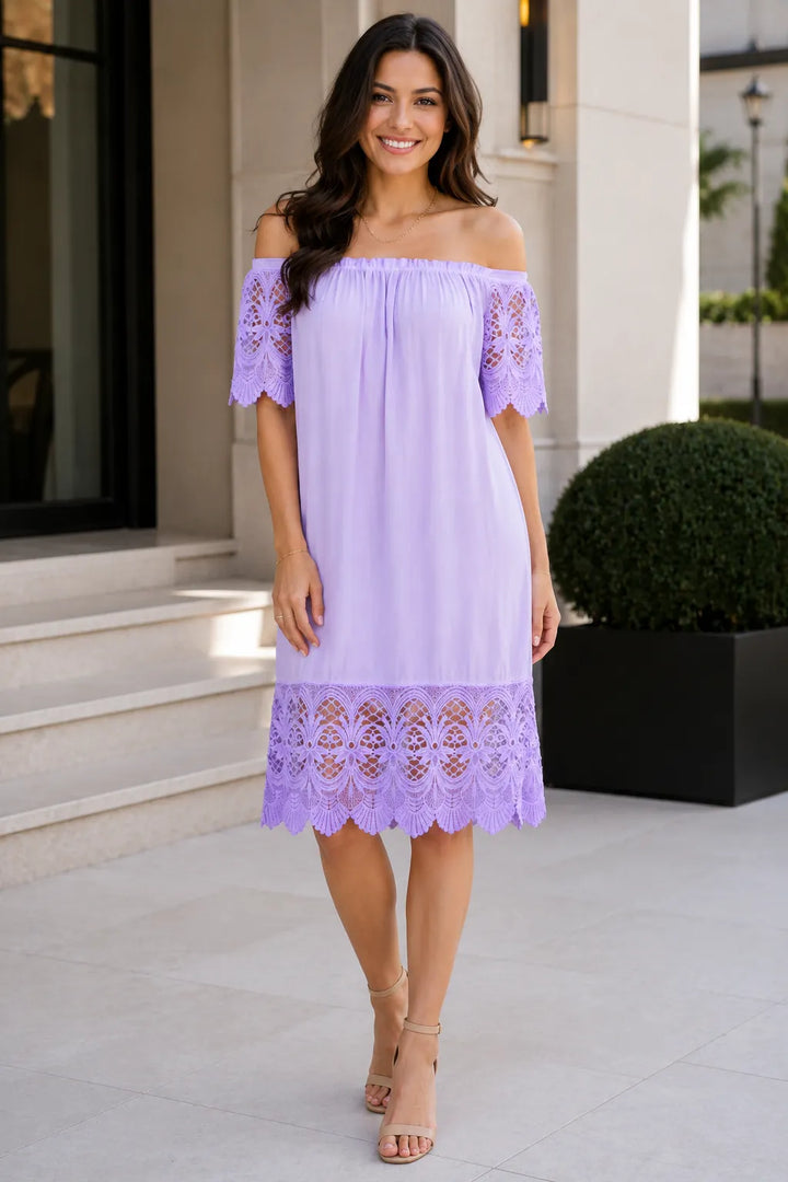 A woman modeling a light purple off-the-shoulder midi dress with lace trim on the sleeves and hem, standing outdoors in front of a building entrance.