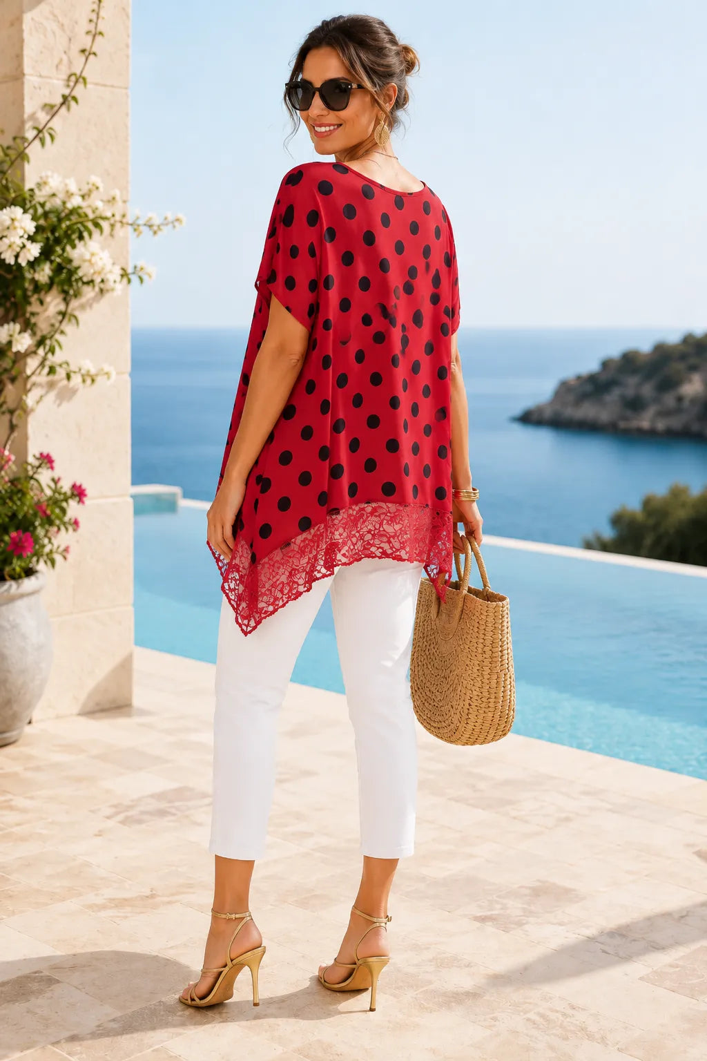 Woman in a red polka dot dress with white pants standing by a pool with ocean view.