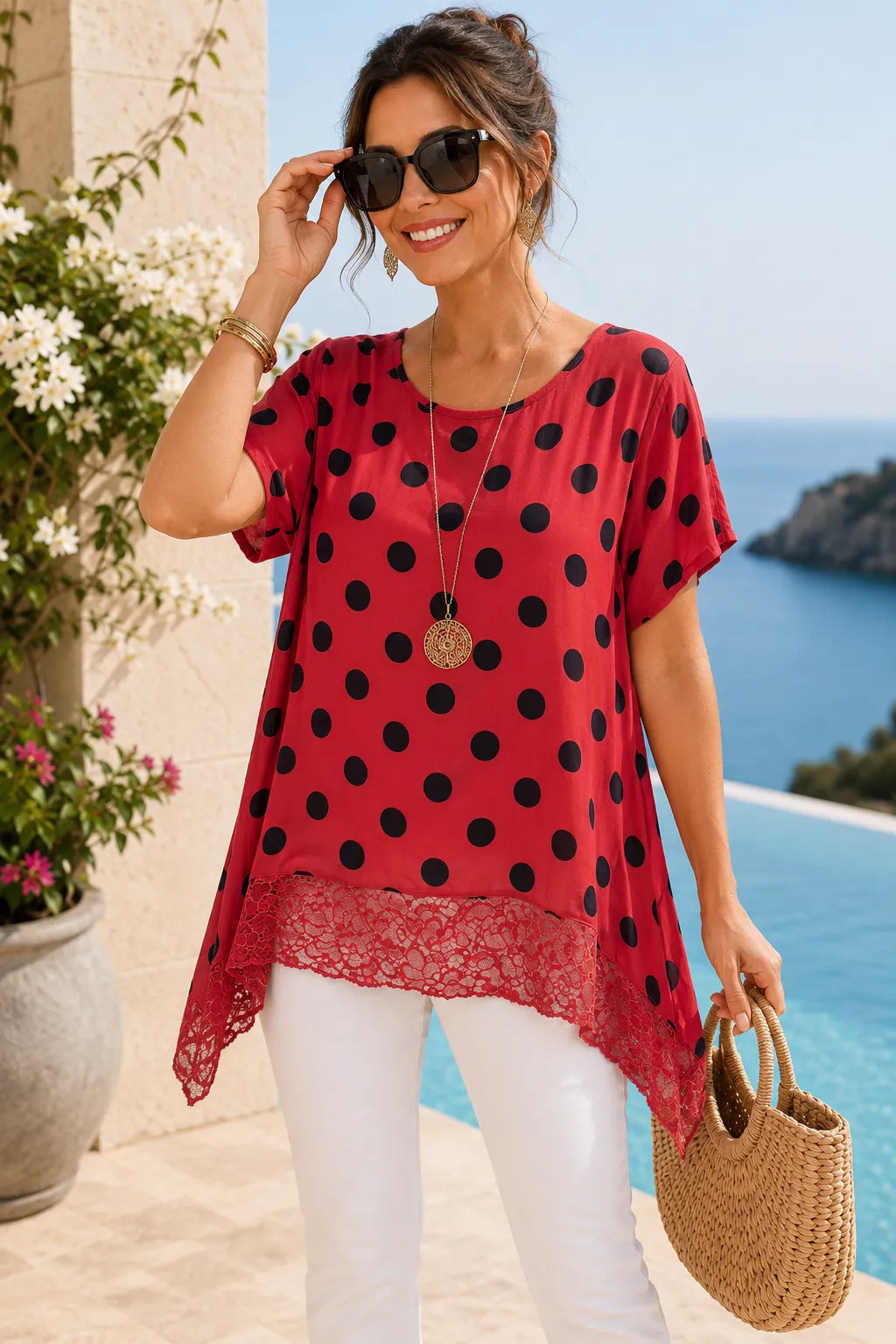 Woman in a red polka dot top and white pants standing by a pool with ocean view.