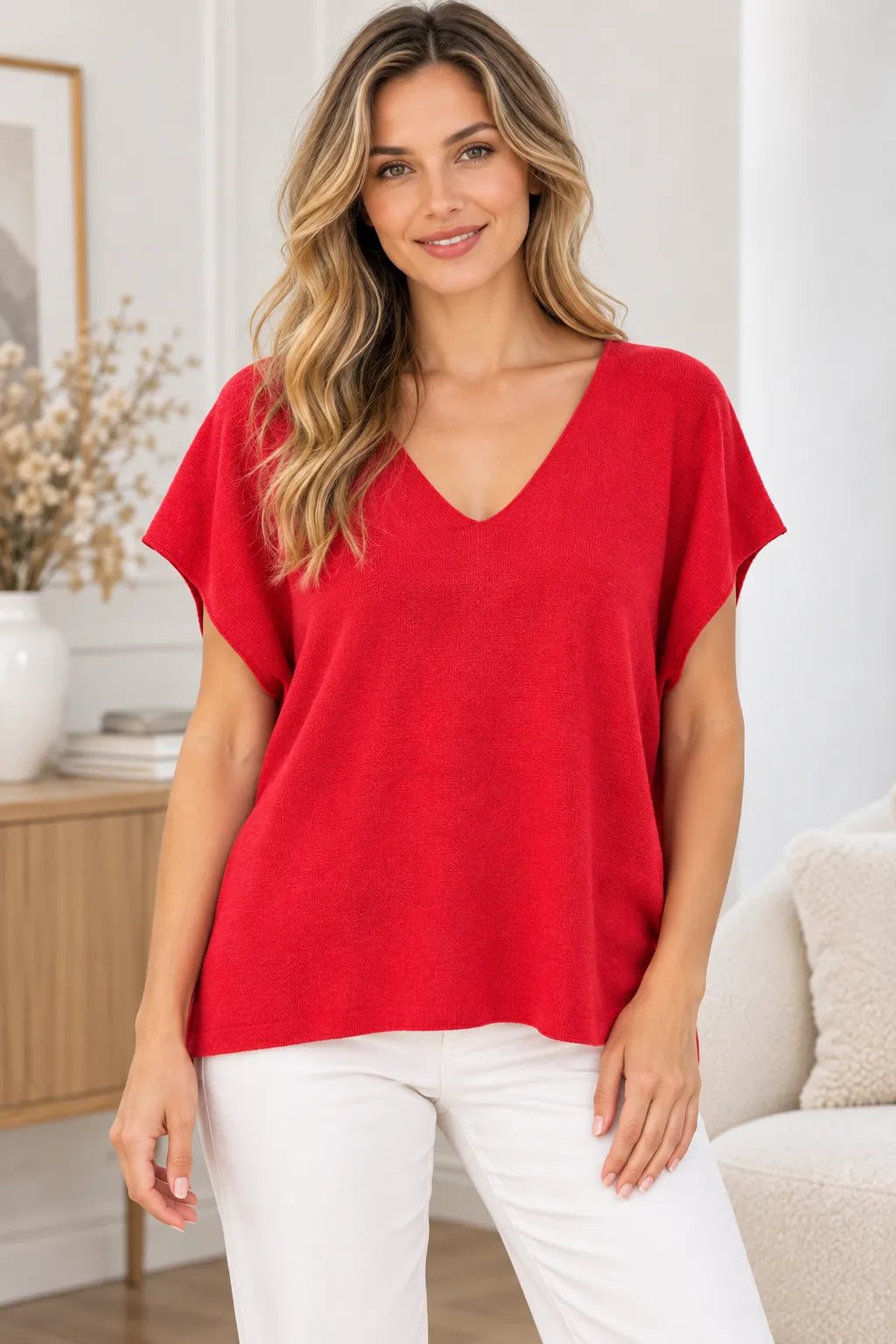 A woman wearing a bright red short-sleeve V-neck top and white pants, standing in a bright, modern living room setting.