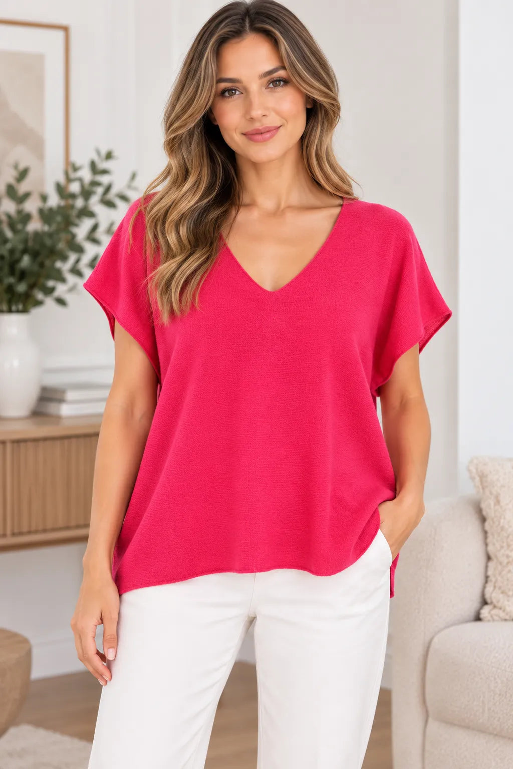 A woman wearing a bright pink short-sleeve V-neck top and white pants, standing in a bright, modern living room setting.