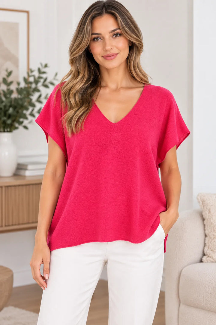 A woman wearing a bright pink short-sleeve V-neck top and white pants, standing in a bright, modern living room setting.