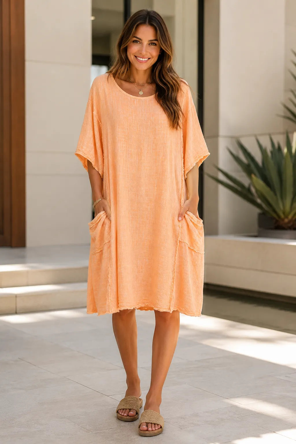 A woman wearing a loose, peach-colored short-sleeve tunic dress with side pockets, standing in a modern outdoor setting with light-colored stone walls and a potted plant.