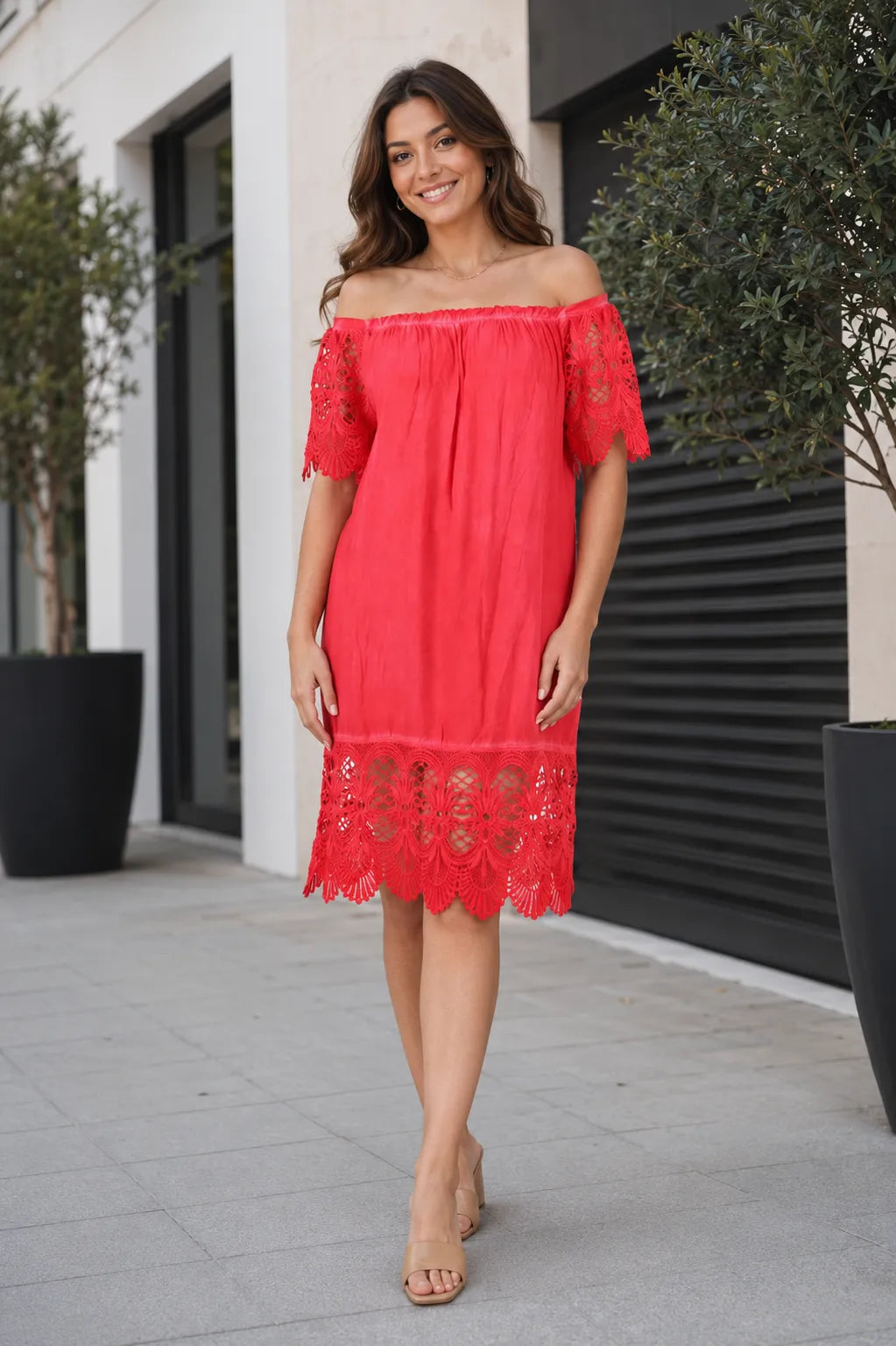 A woman modeling a bright coral red off-the-shoulder lace dress with ruffled sleeves and a scalloped lace hem, standing outdoors on a paved walkway.