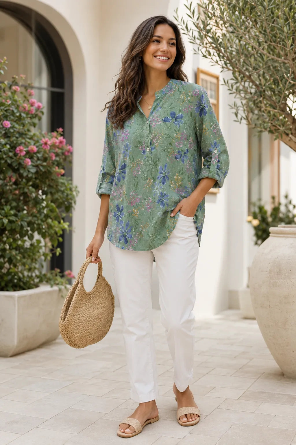 Woman wearing a floral blouse and white pants, standing outdoors with plants and a building in the background.
