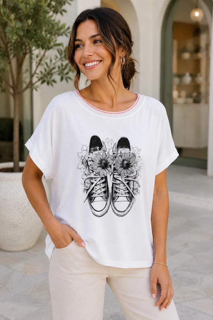 Woman wearing a white t-shirt with a graphic design of sneakers and flowers, standing outdoors.