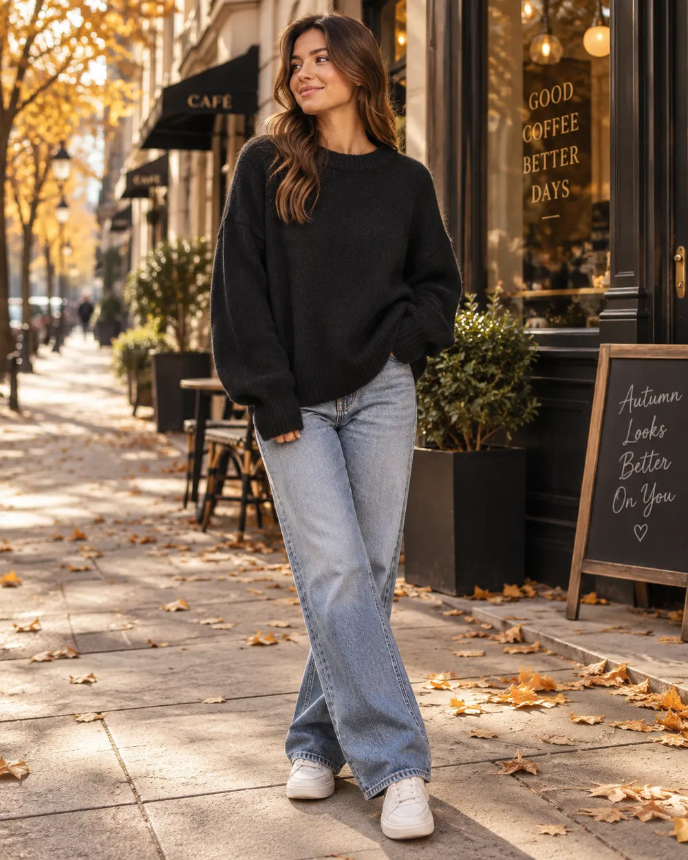 A woman stands on a city sidewalk wearing a black knit crewneck sweater, light-wash blue jeans, and white sneakers. The background shows a storefront with autumn leaves scattered on the pavement.