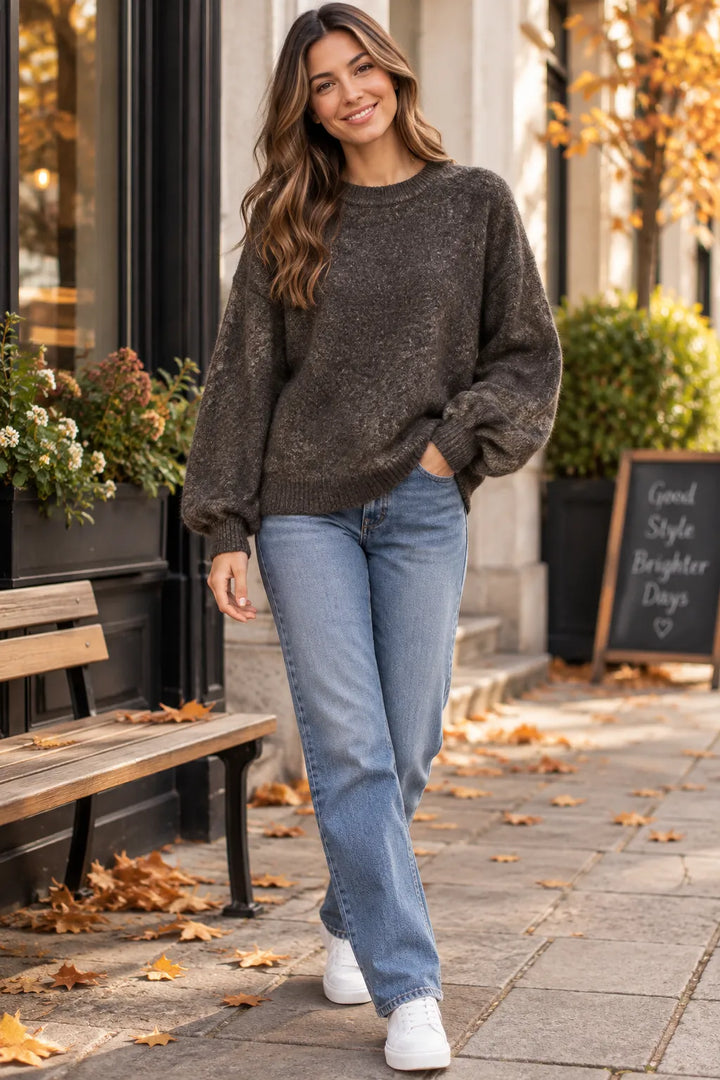 A woman walking outdoors on a city sidewalk wearing a dark brown fuzzy knit sweater, light blue jeans, and white sneakers. The background shows a storefront, bench, potted plants, and fallen autumn leaves.