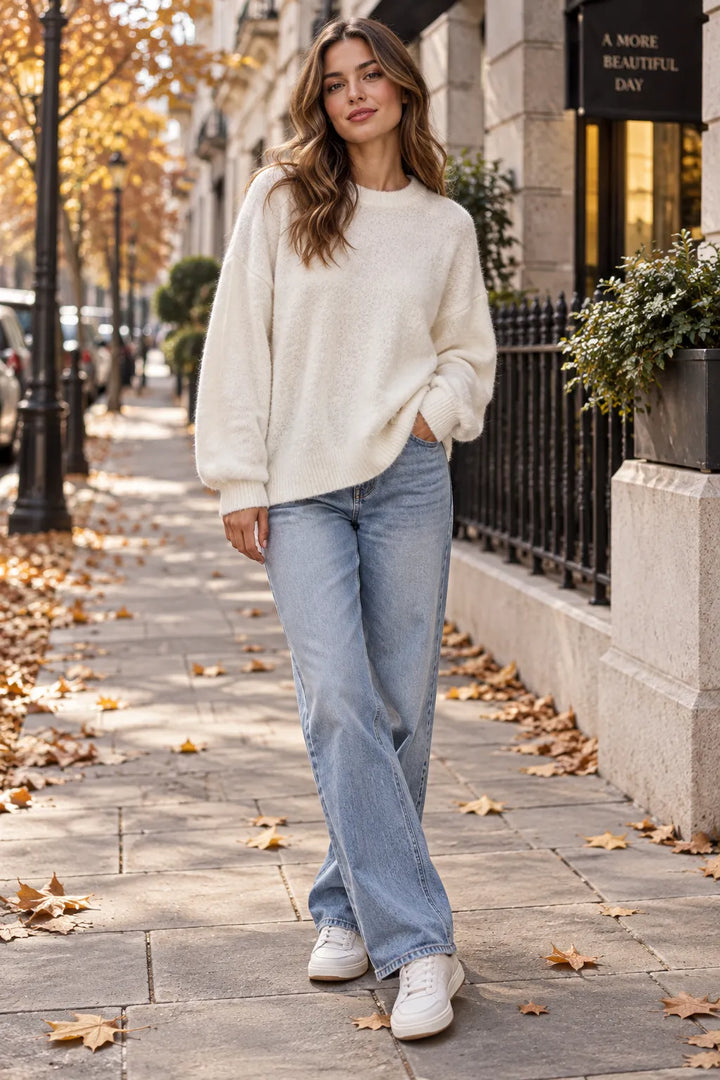 A woman stands on a city sidewalk wearing a cream knit crewneck sweater, light-wash wide-leg jeans, and white sneakers. Fallen autumn leaves are scattered on the pavement, and a stone building with a black wrought-iron fence is in the background.