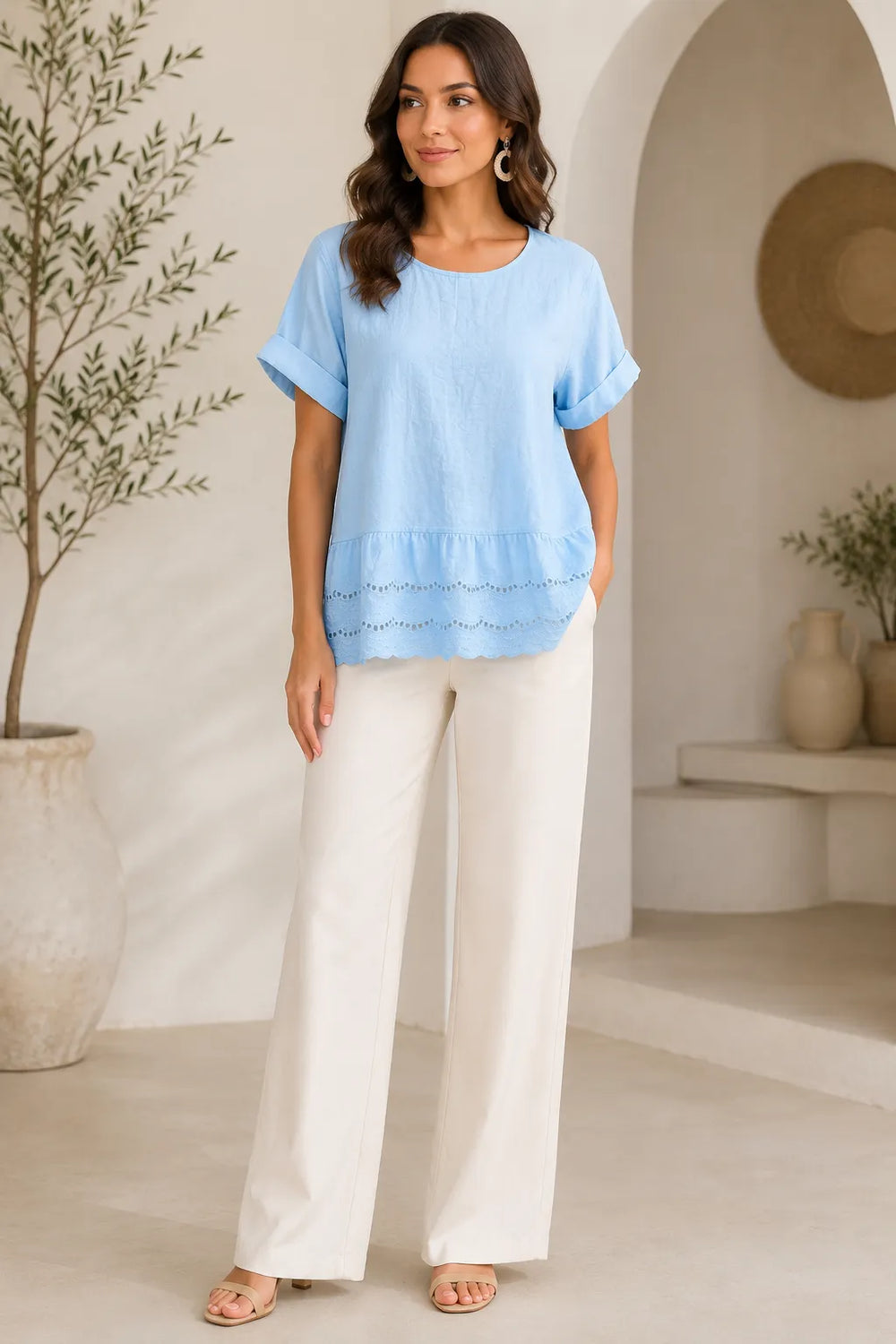 A woman stands in a bright, minimalist indoor setting wearing a light blue short-sleeve top with a scalloped hem and white wide-leg pants.