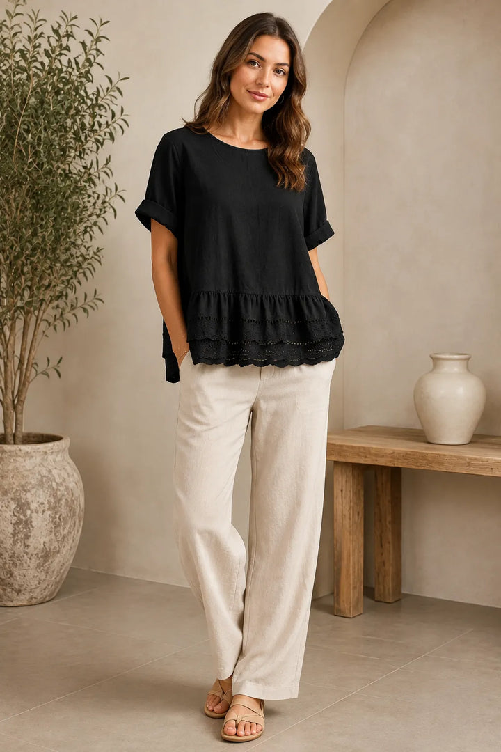 A woman modeling a black short-sleeve peplum top with ruffle trim and cream wide-leg linen pants in a warm, neutral-toned indoor setting.