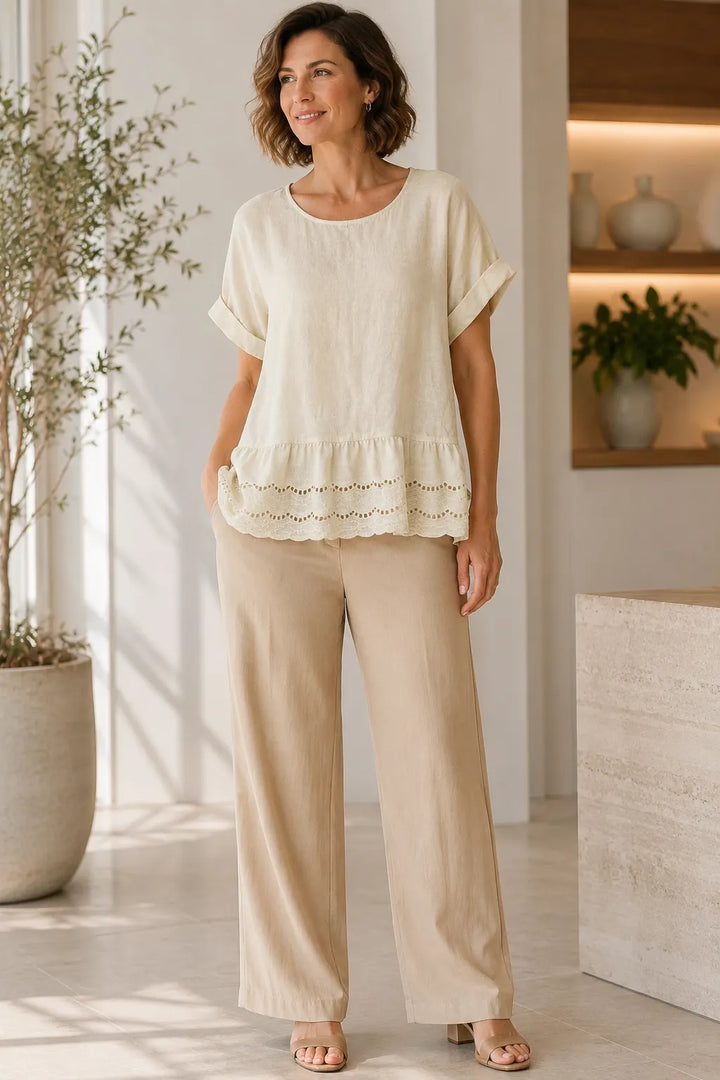 A woman modeling a cream short-sleeve top with a ruffled hem and matching beige wide-leg pants in a bright, modern indoor setting.