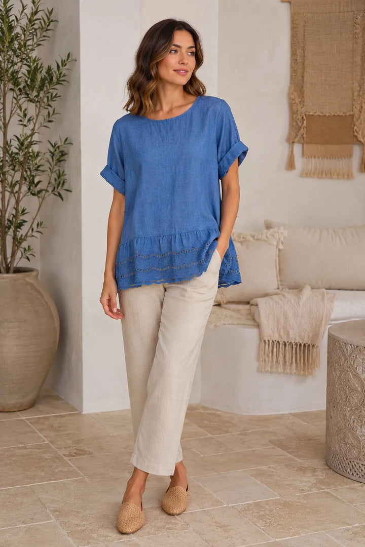 A woman modeling a blue short-sleeve top with a ruffled hem and beige cropped pants in a bright, modern indoor setting with a sofa and potted plant.