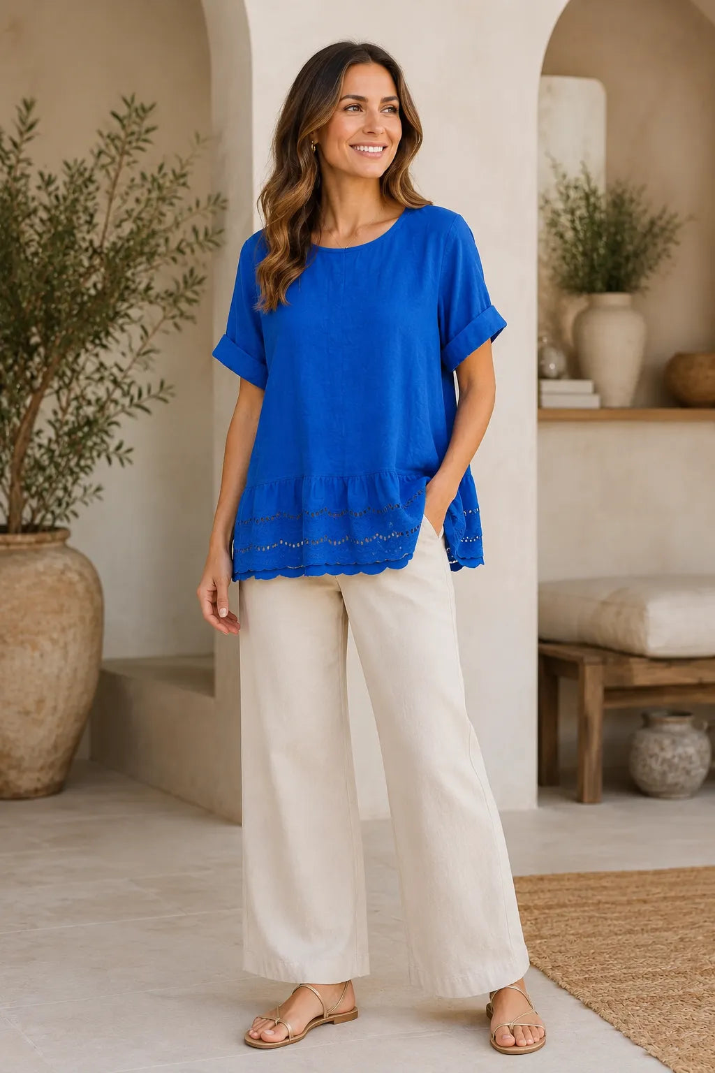A woman modeling a bright royal blue short-sleeve top with a ruffled hem and cream wide-leg pants in a warm, sunlit indoor setting with arches and potted plants.
