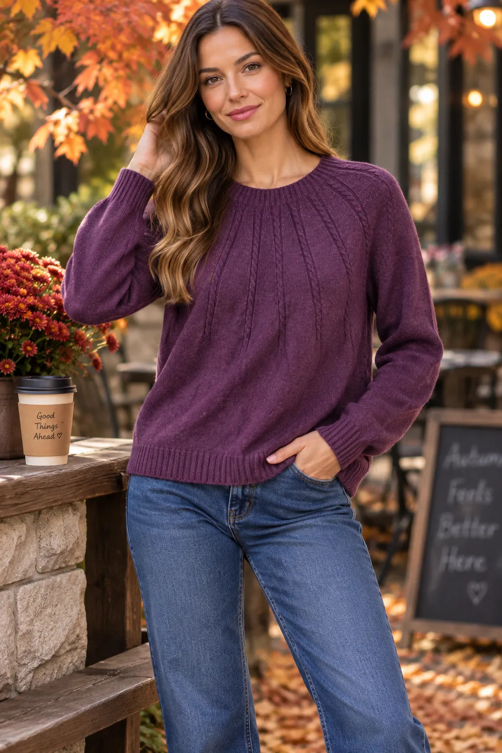 A woman wearing a purple cable-knit crewneck sweater and blue jeans, standing outdoors in an autumn setting with fallen leaves and a coffee cup nearby.