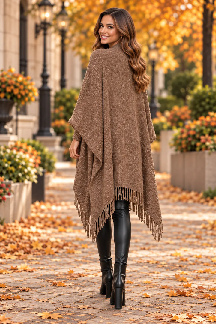 Model smiling over her shoulder wearing a mocha brown knit poncho with a flowing back, relaxed draped fit and fringed hem.