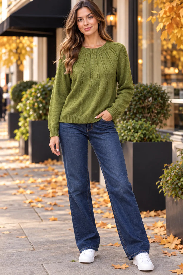 A woman wearing an olive green cable-knit sweater, blue jeans, and white sneakers stands on a city sidewalk with autumn leaves and planters in the background.
